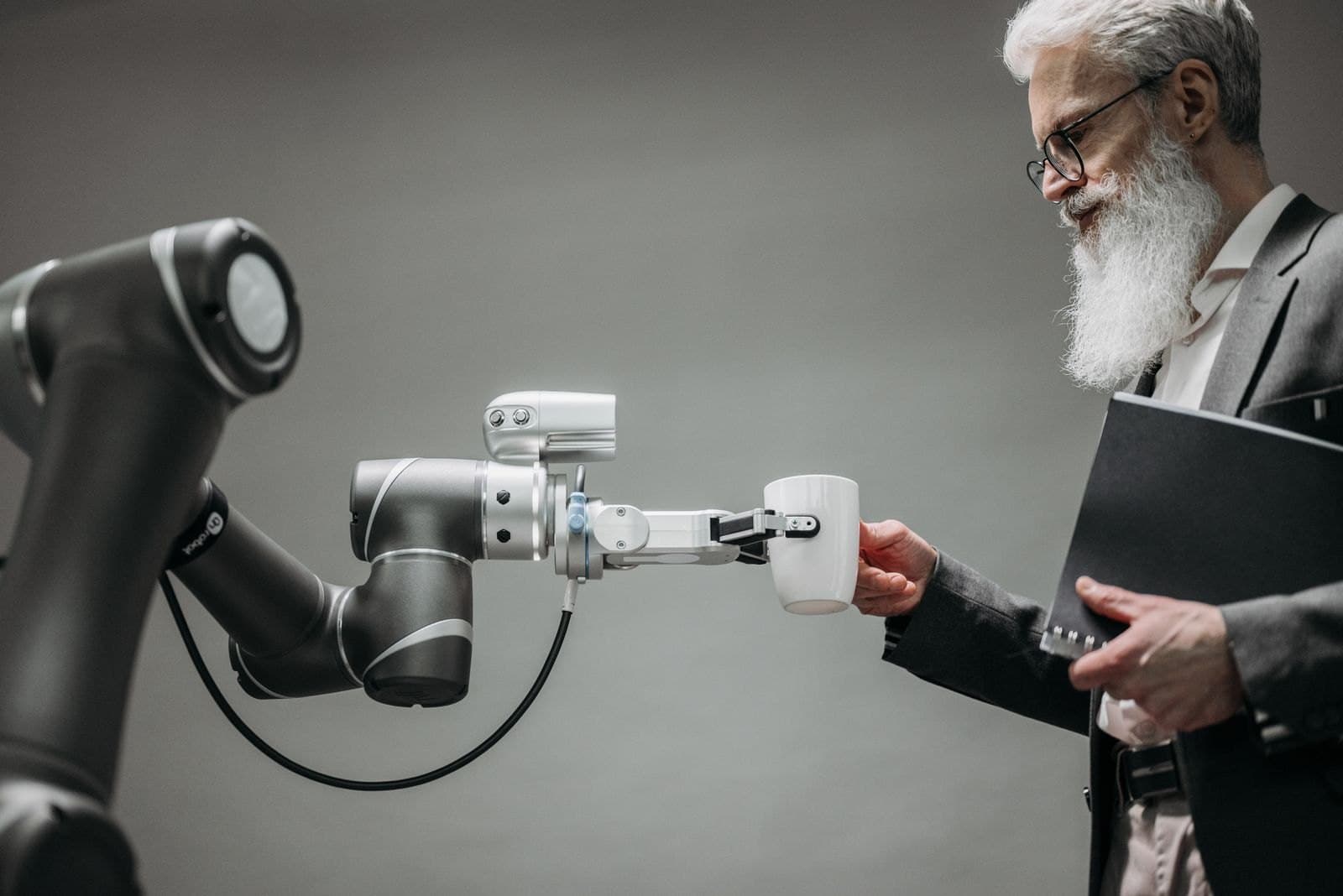A grey robot arm holds out a white mug by its handle to a grey-bearded man in glasses and a dark suit, who takes it while holding a black folder.