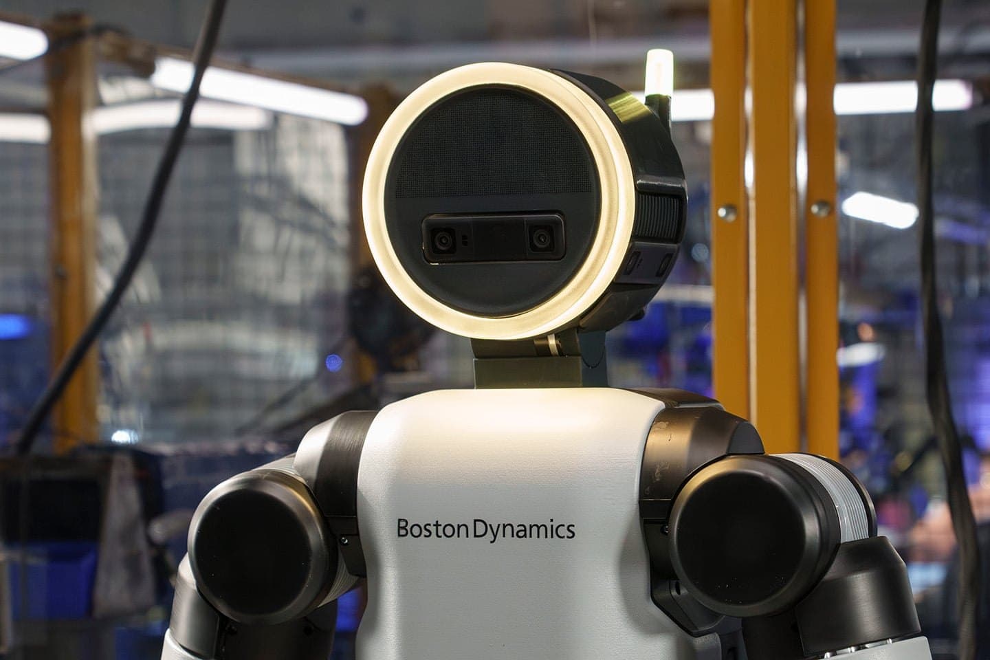 Close-up of Boston Dynamics' Atlas humanoid robot: a round head ringed with white light above a white torso carrying the company's name, in a lab with yellow safety posts.