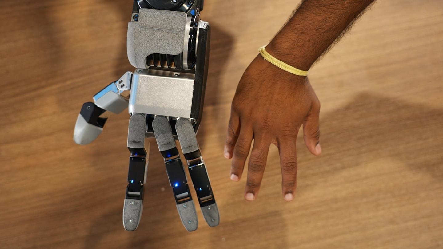 Boston Dynamics' four-fingered GR3 robot hand laid flat on a wooden table next to a person's hand, the two about the same size.