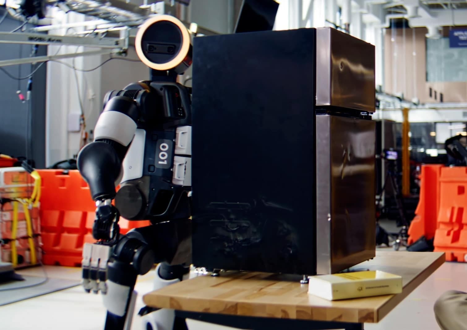 An Atlas humanoid robot numbered 001 gripping a black mini-fridge that rests on a wooden workbench in Boston Dynamics' lab.