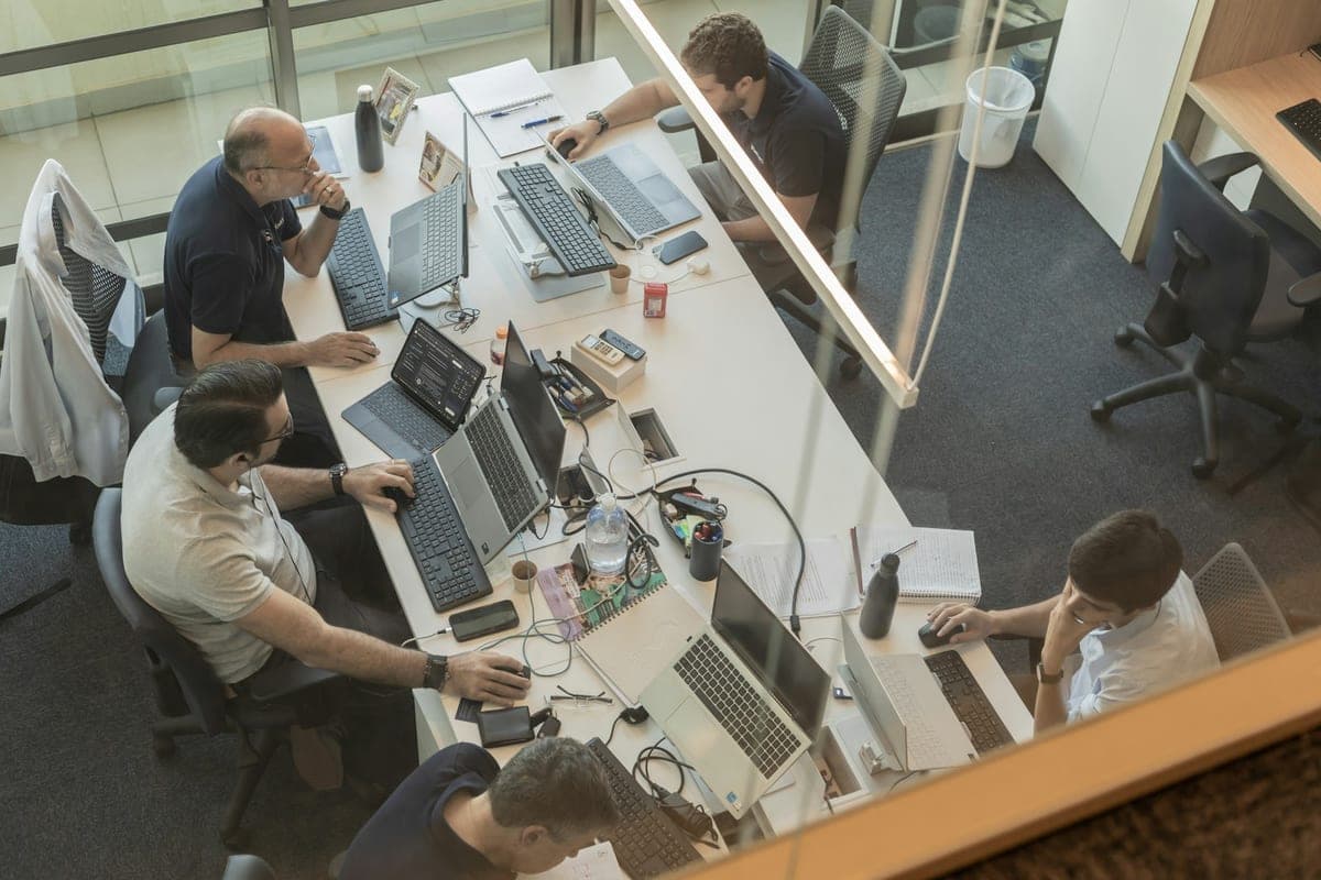 Four people working side by side at laptops and monitors in a shared office, seen from above.