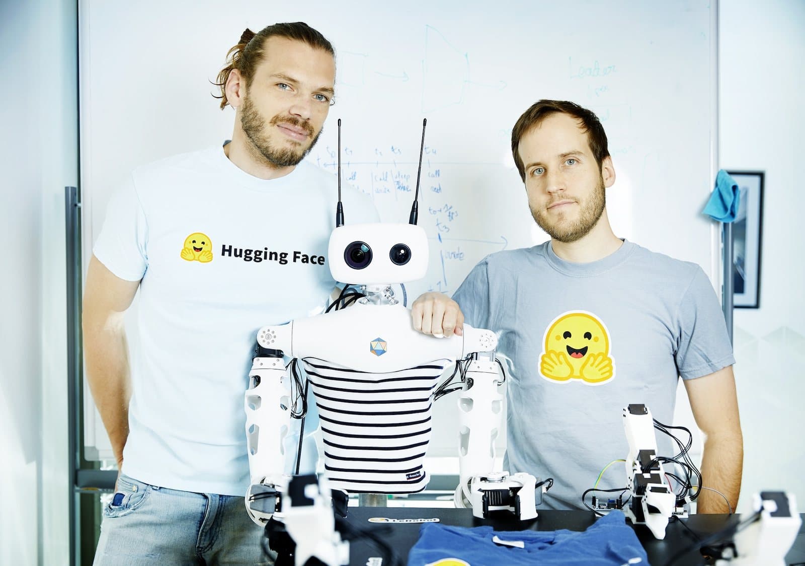 Two members of Hugging Face's LeRobot team in Hugging Face T-shirts standing in front of a whiteboard on either side of a white Reachy humanoid robot in a striped top.