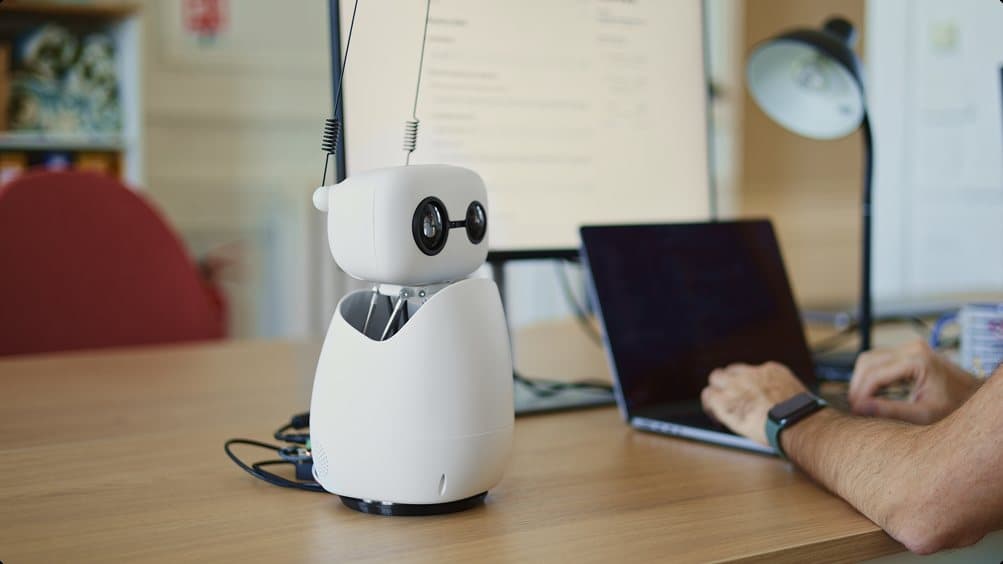 A small white Reachy Mini robot with two antennas and round camera eyes on a wooden desk, next to a person typing on a laptop.