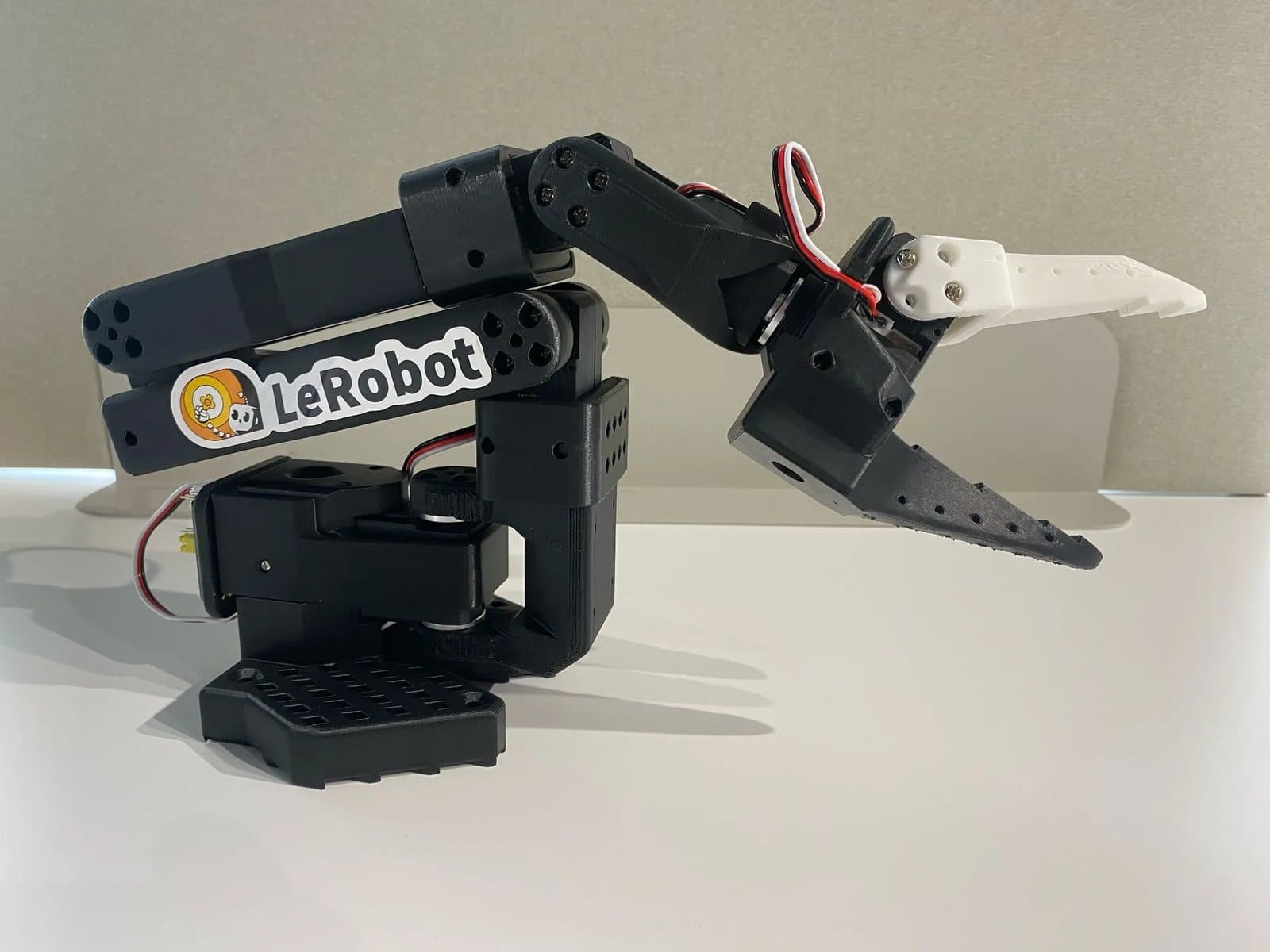 A black 3D-printed SO-101 robot arm with a LeRobot sticker and a white gripper tip, on a white desk.