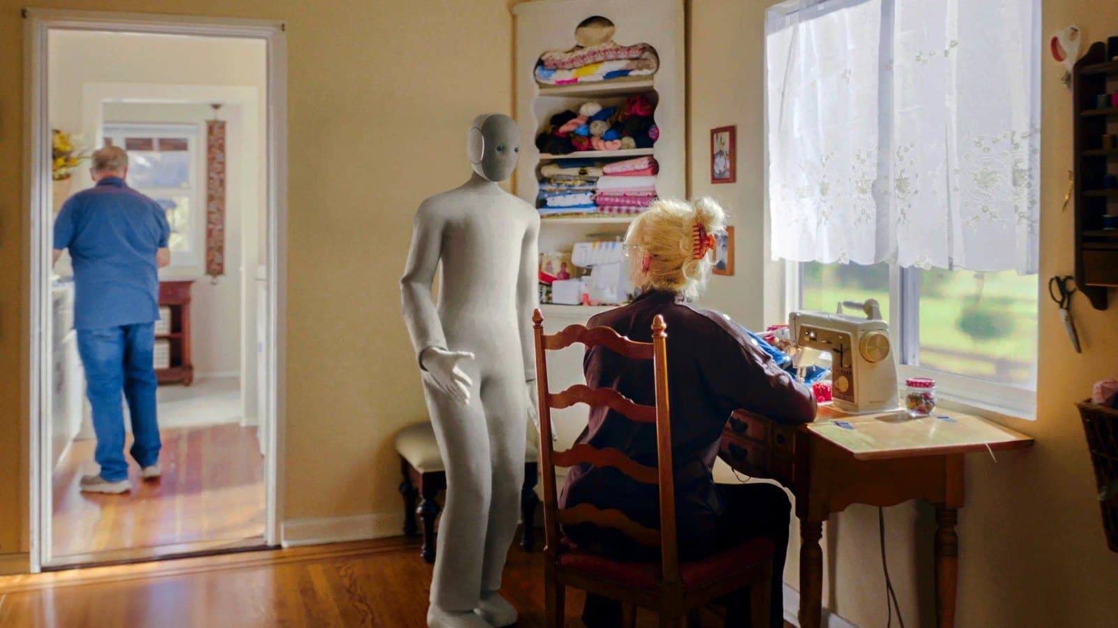 The NEO humanoid robot standing in a sunlit sewing room beside a woman seated at a sewing machine, with a man walking through the doorway behind.