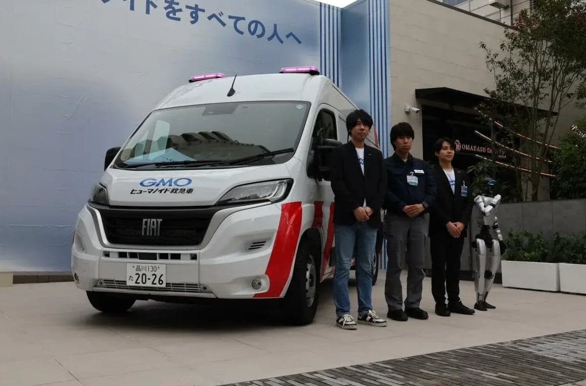 GMO Air's white humanoid ambulance van with red stripes and roof lights, three engineers and a small humanoid robot standing beside it outside GMO's Tokyo office.