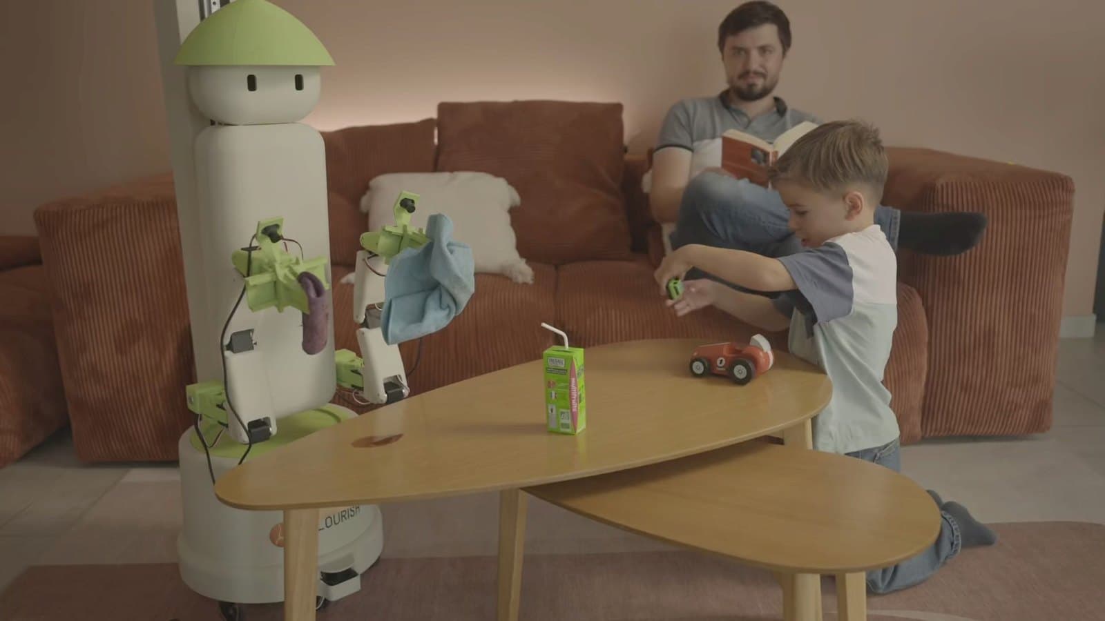 The Flourish One home robot, white with a green hat-shaped head, wiping a coffee table while a father reads to his child on the sofa.