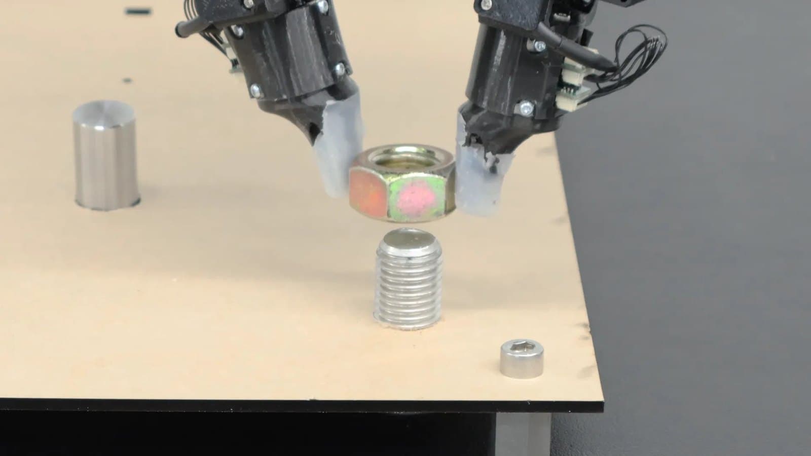 Two robot fingertips with touch sensors holding a metal nut above a threaded bolt on a workbench.