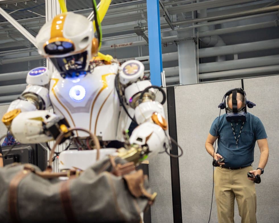 NASA's white and gold Valkyrie humanoid robot holding a cargo bag while team lead Shaun Azimi operates it wearing a headset.