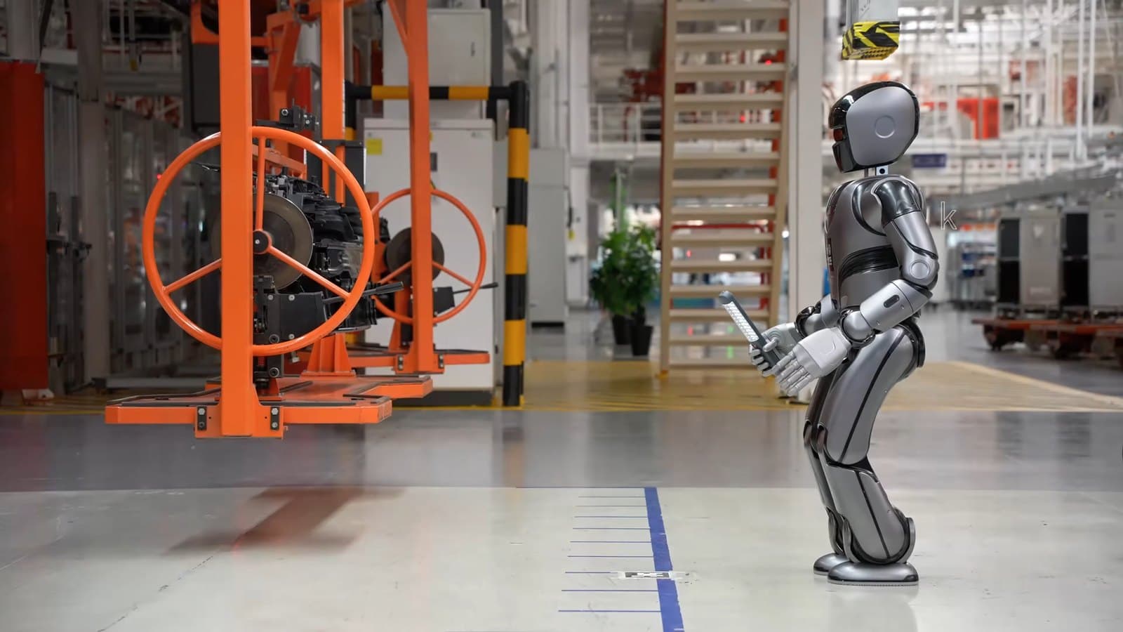 A silver and black UBTECH Walker S Lite humanoid robot standing on a car plant floor beside orange overhead assembly equipment.
