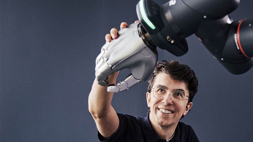 A smiling man in glasses pressing his palm against a silver five-finger robot hand mounted on a dark robot arm.