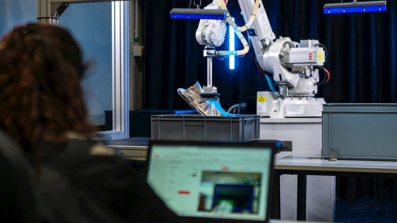 A person at a laptop in the foreground while an ABB robot arm behind them lifts a bagged garment out of a grey tote in a sorting cell lit by blue light bars.