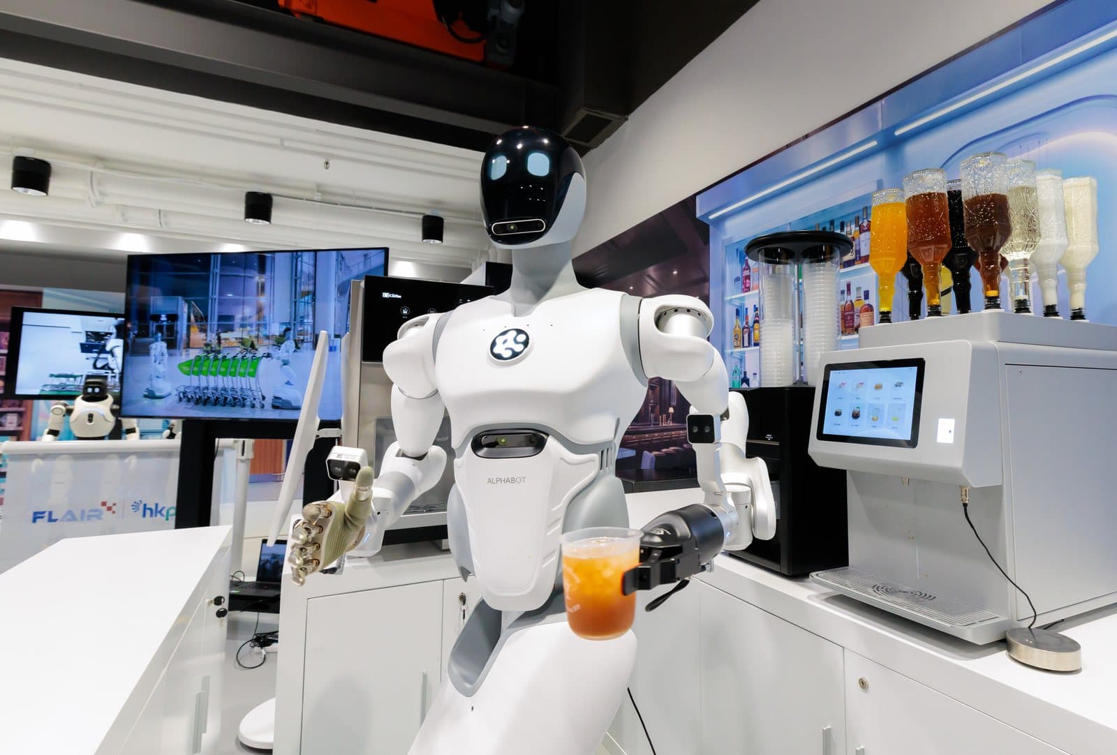 A white AlphaBot 2 humanoid robot holding a cup of iced tea at a drinks counter, with bottles on a lit shelf and FLAIR and HKPC logos on a screen behind it.