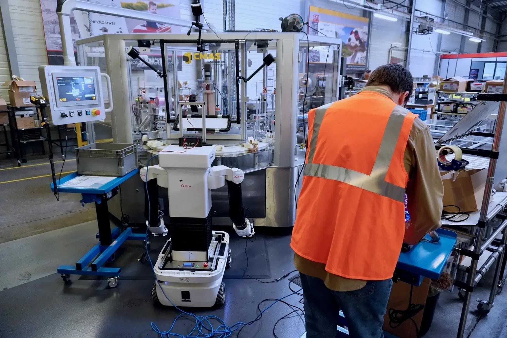 An operator in an orange hi-vis vest works at a packaging cell beside a white wheeled humanoid robot with two arms, in a busy factory hall.