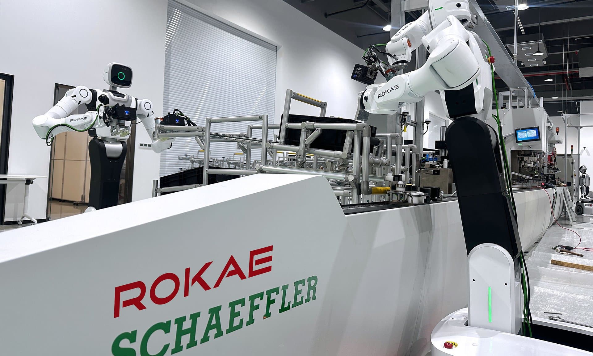 Two white ROKAE wheeled humanoids with force-controlled arms working along an assembly line in a bright factory, behind a white barrier printed with the ROKAE and Schaeffler logos.