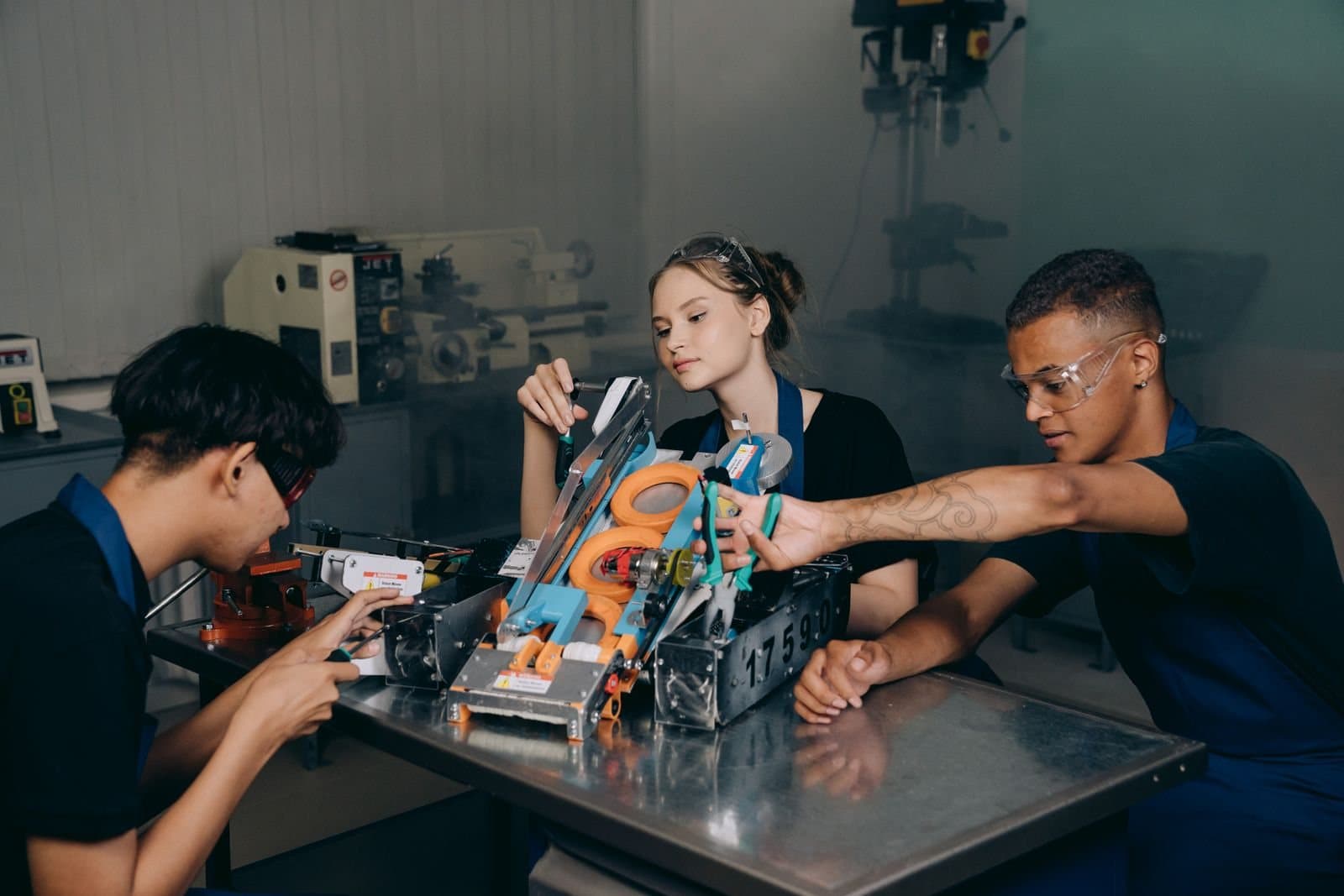 Three students in safety glasses working together on a small robot built from metal parts, on a steel workbench in a workshop.