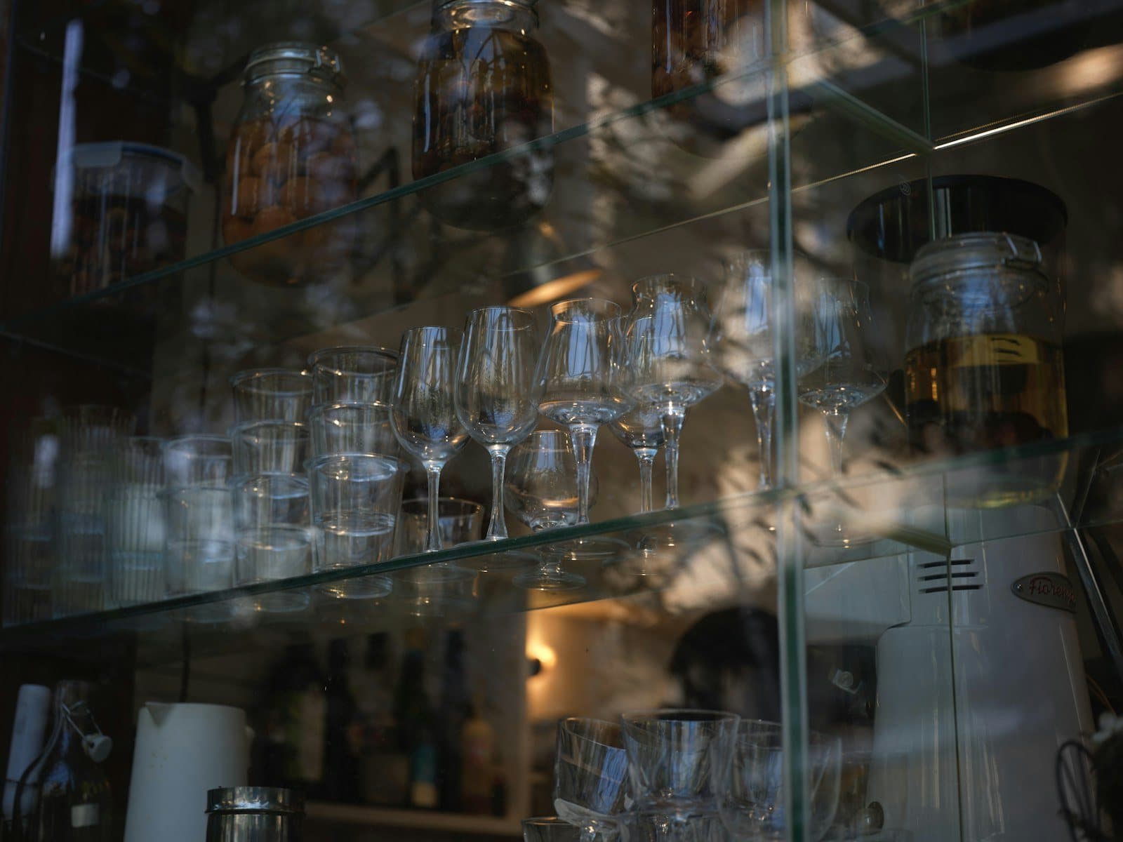 Clear wine glasses and glass jars on glass shelves inside a dimly lit cabinet.