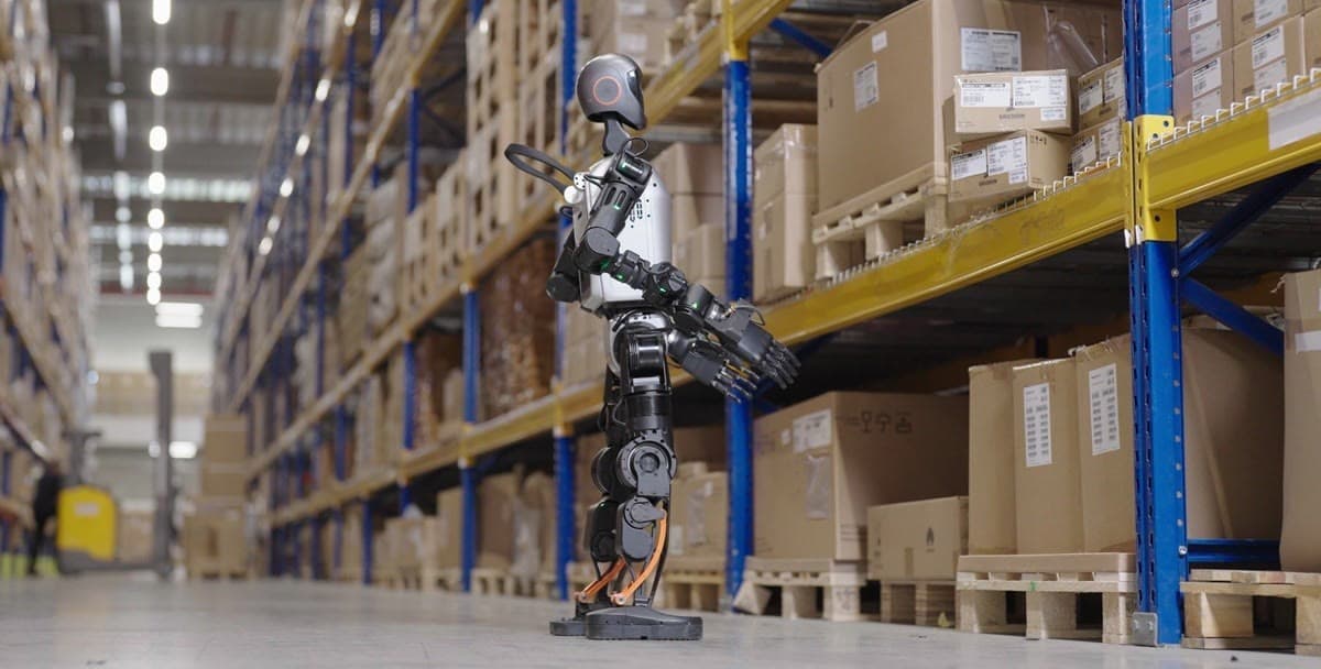 A humanoid robot standing in a warehouse aisle, inspecting stacked boxes on pallet racking.
