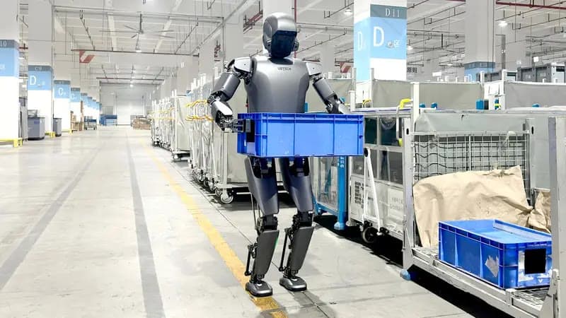 A UBTech humanoid robot carrying a blue parts tote along a factory aisle lined with trolleys.