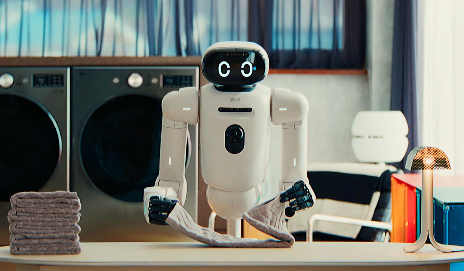 LG's CLOiD home robot folding a towel on a table in front of a washer and dryer, beside a stack of folded laundry.
