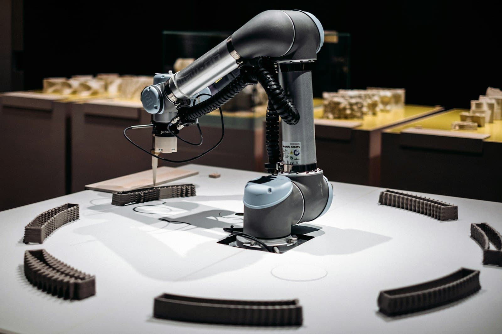 A collaborative robot arm placing a small piece on a white table, with rows of finished pieces around it.