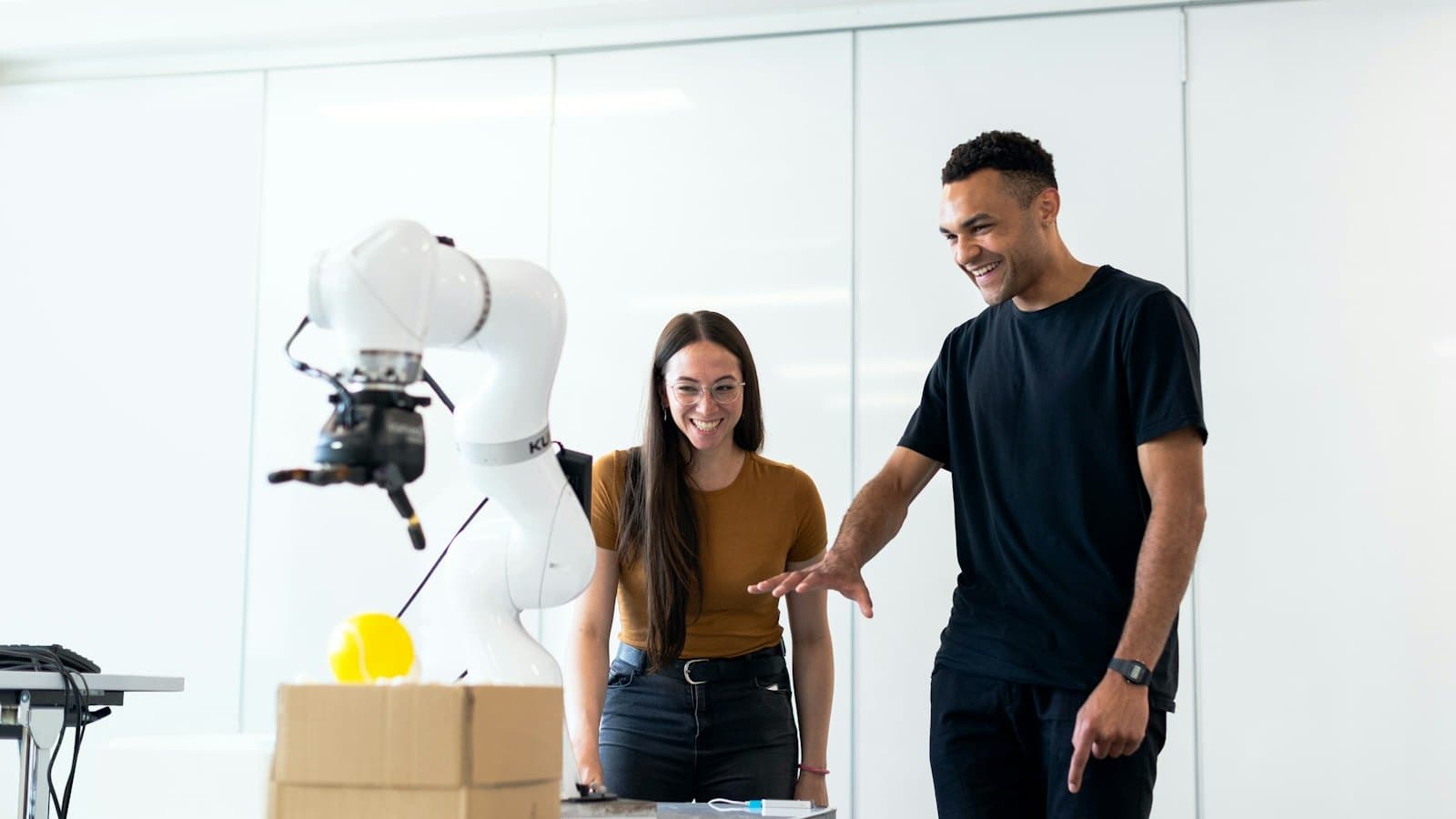 Two engineers standing beside a white robot arm whose gripper hovers over a yellow ball on a cardboard box.