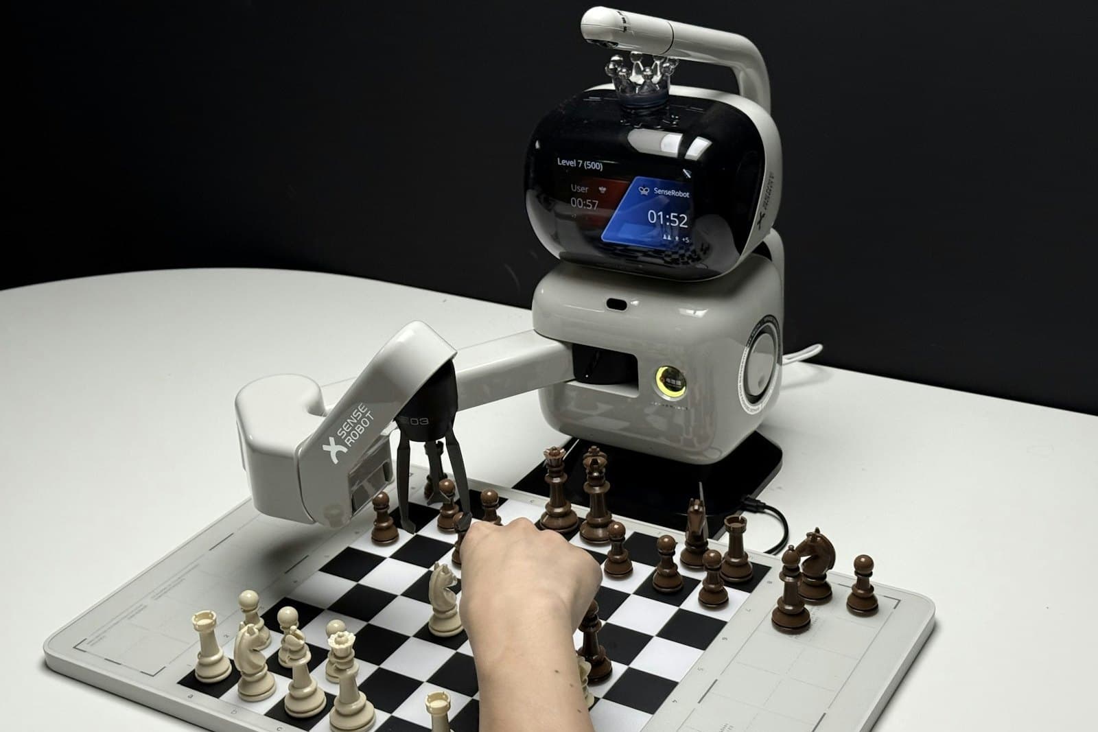 A small desktop robot arm moving a chess piece on a board while a human hand reaches for another piece.