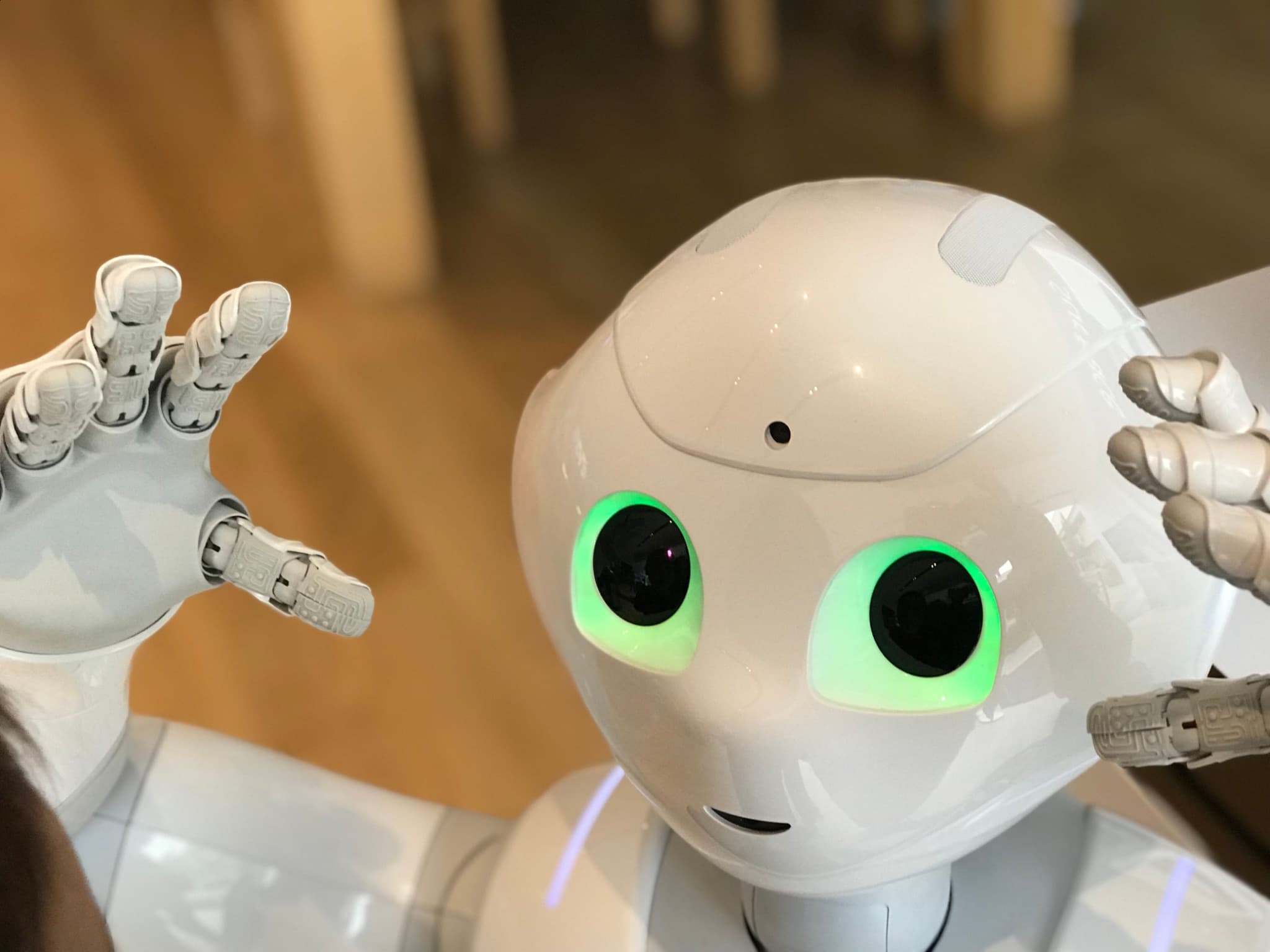 A friendly white humanoid robot with glowing green eyes raising both hands.